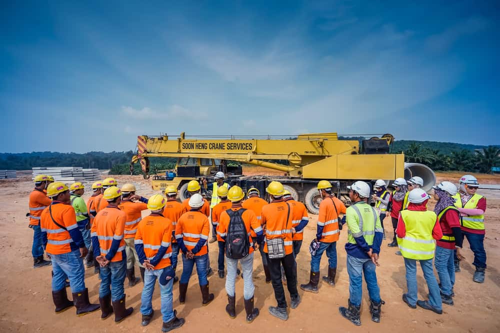 a group of people in orange vests and helmets standing in front of a large yellow crane doing personnel training